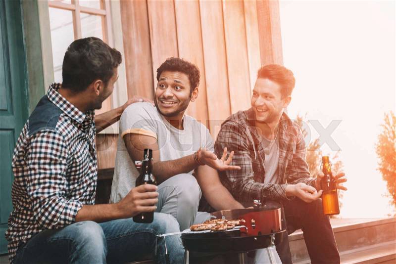Smiling young friends drinking beer and talking while making barbecue , stock photo