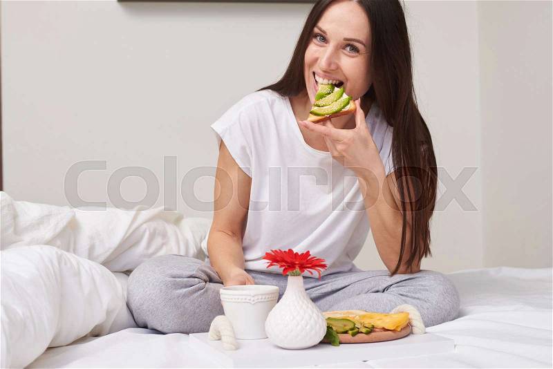 Mid shot of smiling woman in pajamas eating sandwich and enjoying romantic breakfast sitting in the bed with copy space, stock photo