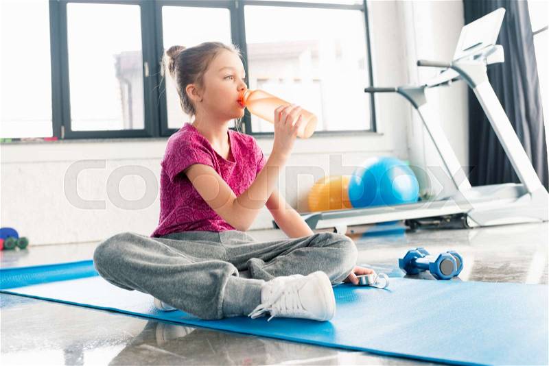 Adorable girl in pink shirt sitting on yoga mat and drinking water in gym , stock photo