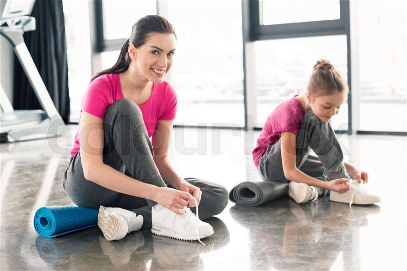 Mother and daughter in pink shirts and sneakers tying shoelaces in gym , stock photo