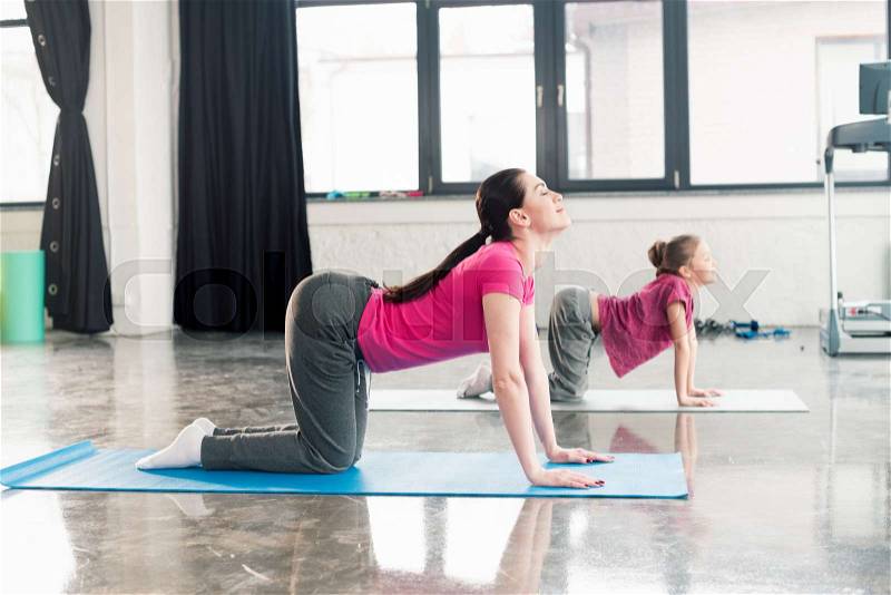 Mother and daughter in pink shirts practicing yoga in Cat pose in gym, stock photo