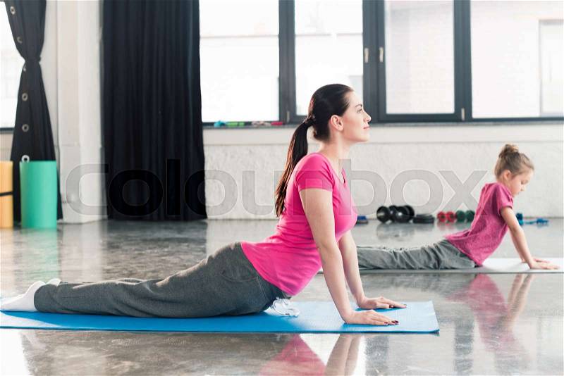 Mother and daughter in pink shirts practicing yoga in Cobra Pose in gym, stock photo