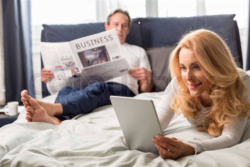 Middle aged woman using digital tablet and lying on bed, man reading newspaper behind, stock photo