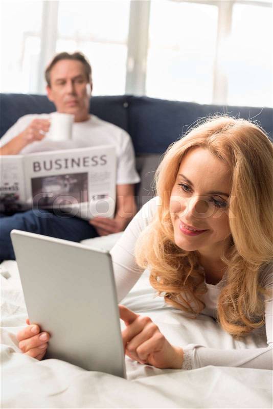 Middle aged woman using digital tablet and lying on bed, man reading newspaper behind, stock photo