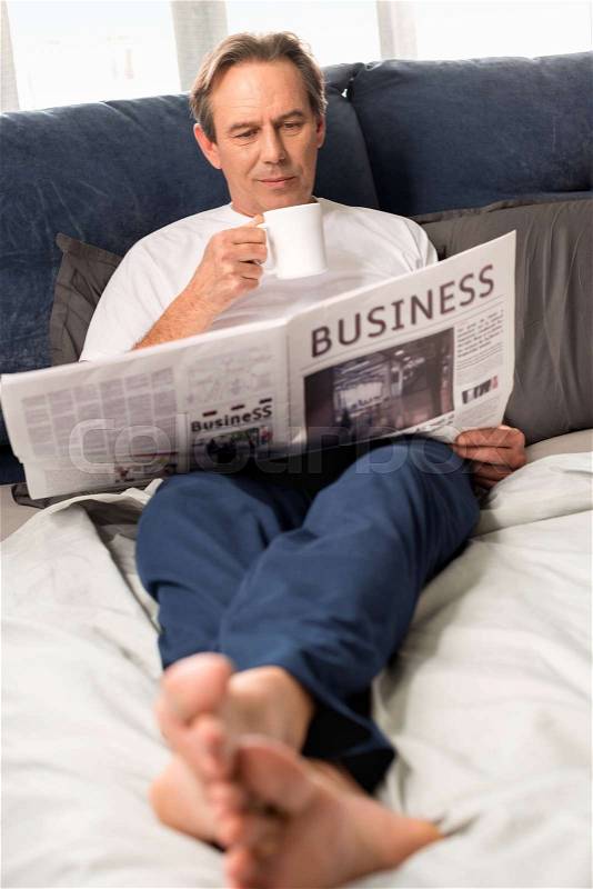 Middle aged man reading newspaper and lying on bed at home , stock photo