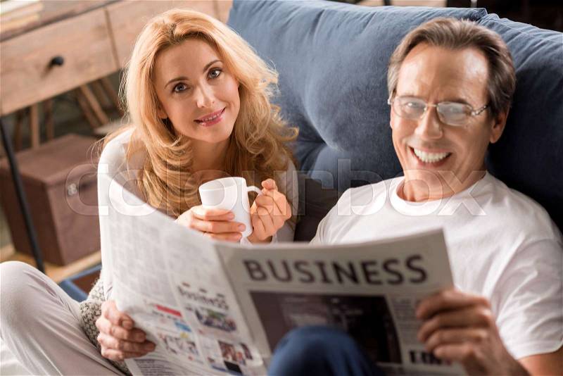 Happy middle aged couple sitting on bed, man reading newspaper at home , stock photo