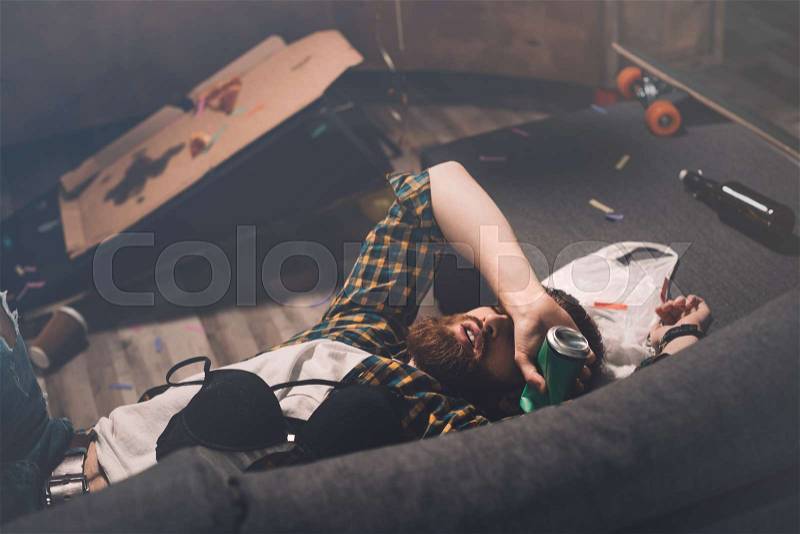 Drunk bearded young man sleeping on couch with bra and beer can after party , stock photo