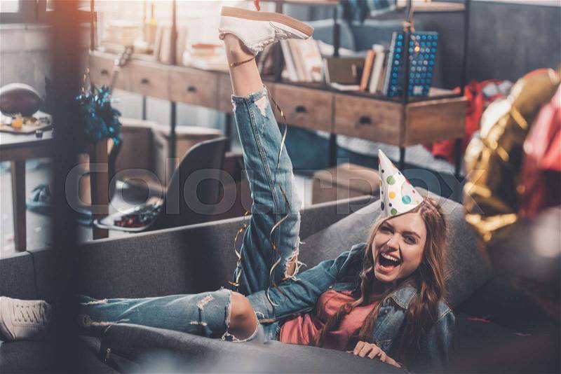 Smiling woman in birthday hat sitting on sofa in messy room after party, stock photo