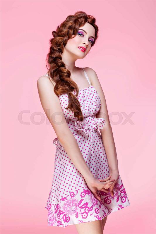 Beautiful young woman with long red braided hair and bright colourful makeup wearing dress standing on pink background. Pin up style. Copy space, stock photo