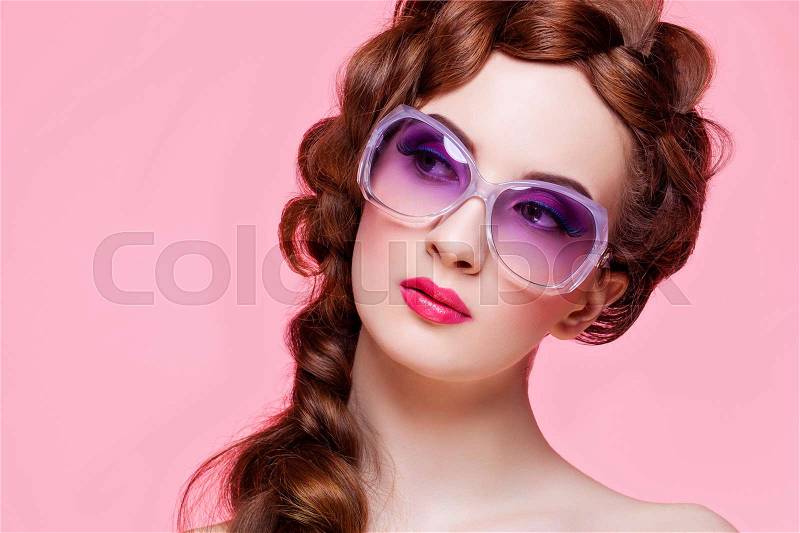 Beautiful young woman with red braided hair, bright make-up and purple sunglasses over pink background. Pin up style. Copy space, stock photo