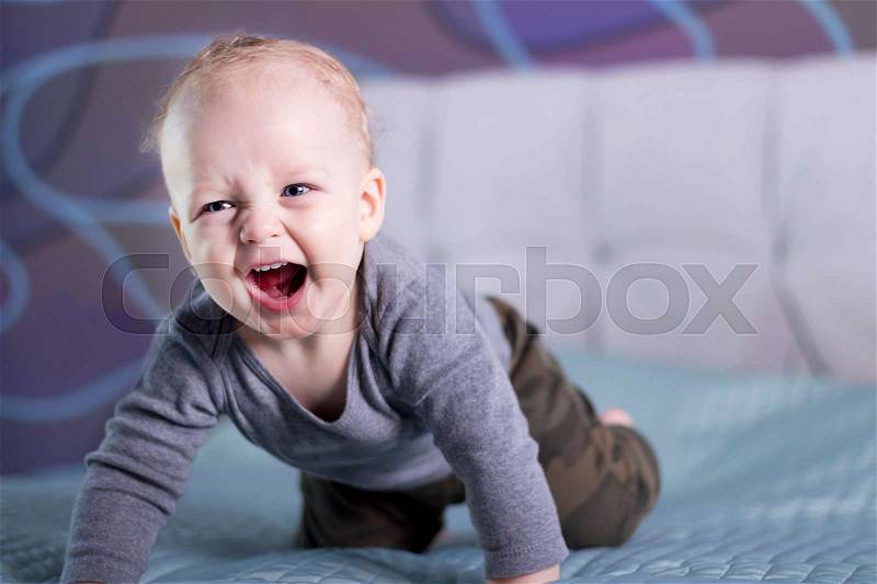 Crying happy baby boy on the bed. Laughing toddler kid looking at camera. Crawling infant in his room, stock photo