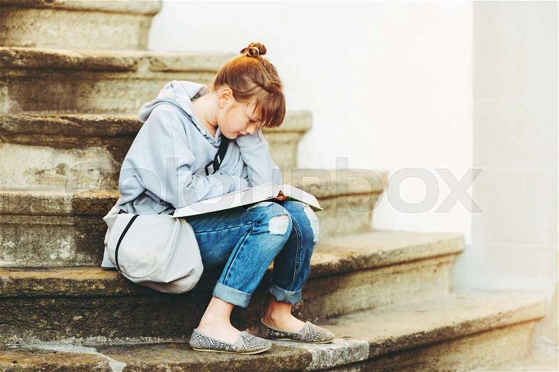 Kid girl reading book outside, sitting on stairs, wearing grey sweatshirt and backpack, stock photo