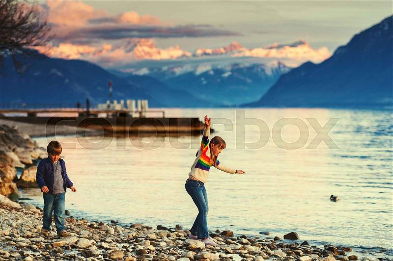Two happy kids, little brother and big sister, playing together by lake Geneva at sunset with swiss mountains Alps on background. Image taken in Lausanne, Switzerland, stock photo
