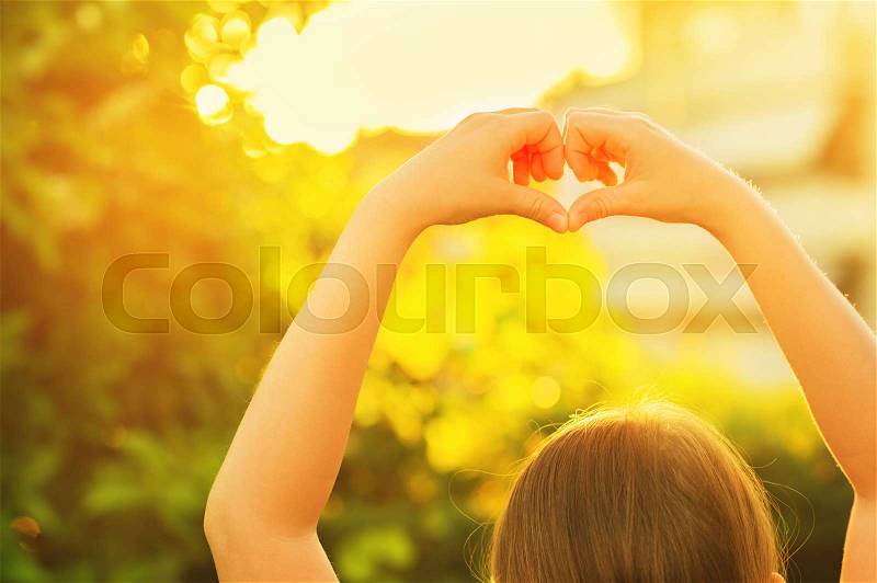 Little girl making heart sign with her hands in the summer garden at sunset, stock photo