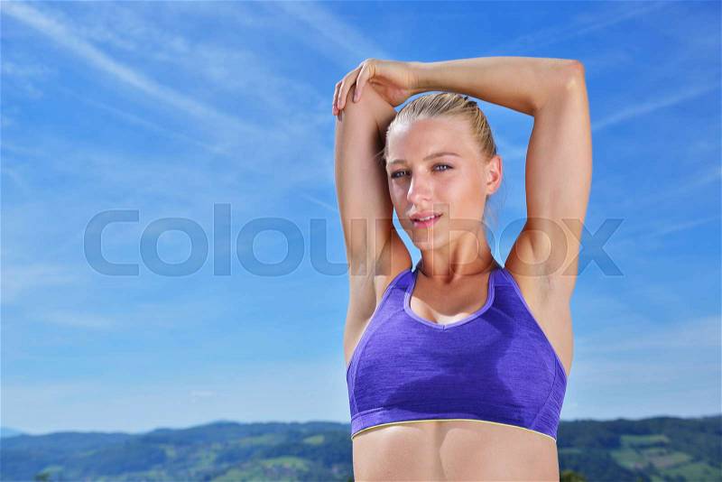 Young sports woman stretching her arms up while exercising on a mountain with a blue sky, stock photo