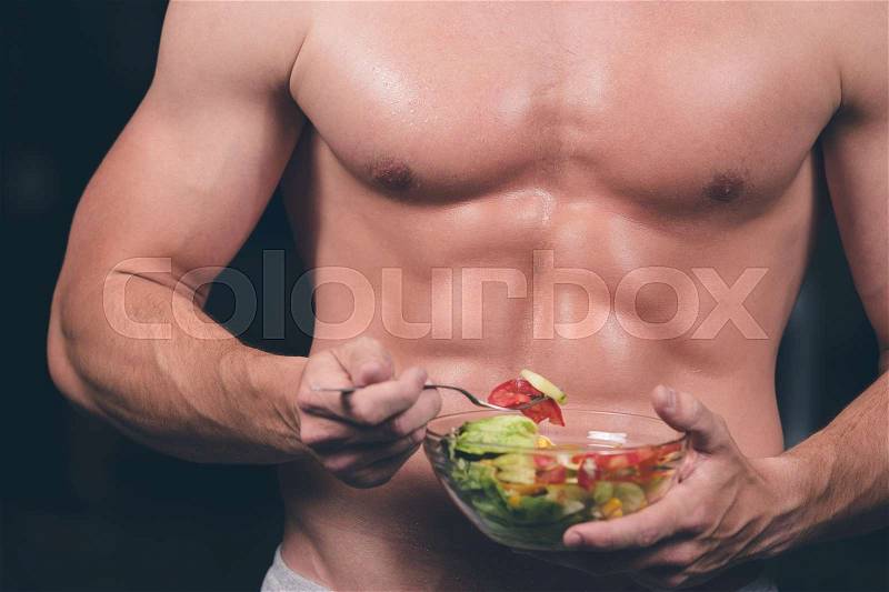 Shaped and healthy body building man holding a fresh salad bowl, shaped abdominal, isolated on dark background, stock photo