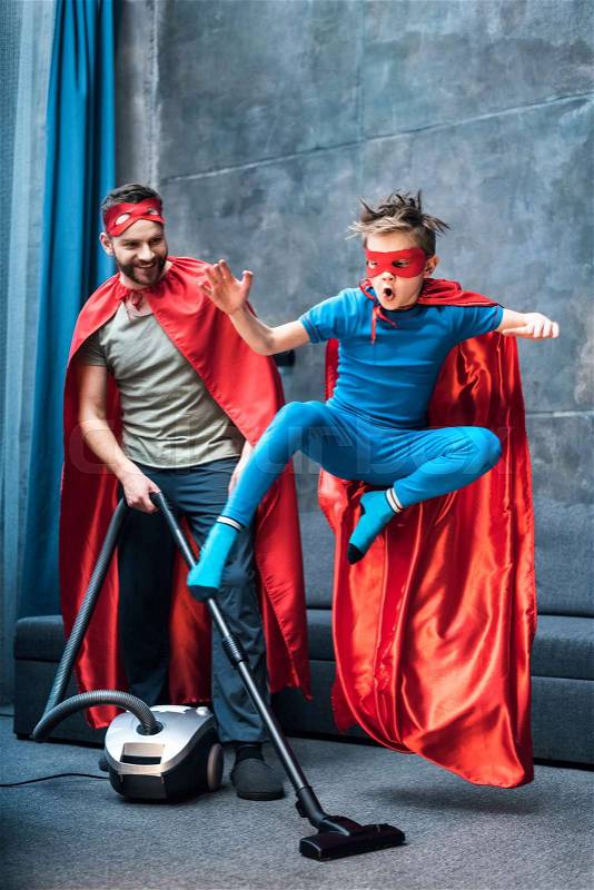 Father in superhero costume vacuuming carpet while son in superhero costume jumping, stock photo