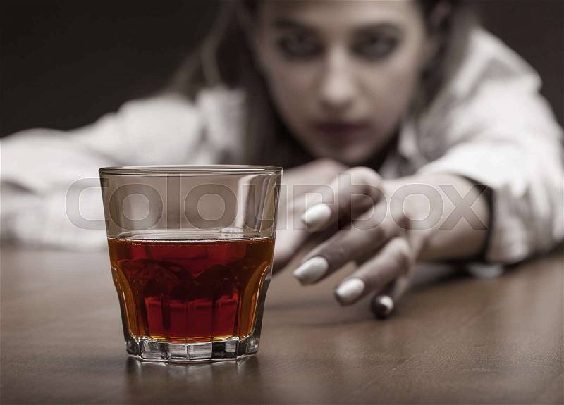 Young woman in depression, drinking alcohol on dark background. Focus on the glass, stock photo