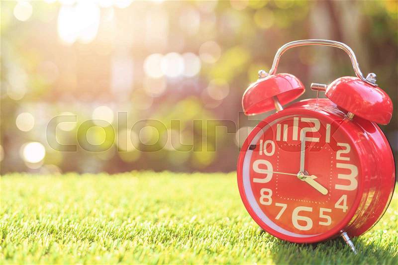Close up red alarm clock on green grass with sunlight blur and bokeh background in morning time, stock photo