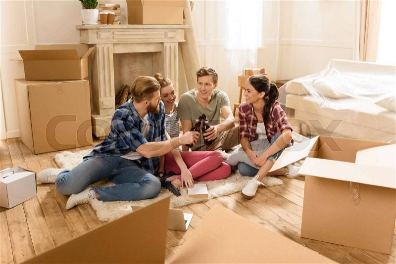 Happy young friends drinking beer while sitting on carpet in new house, stock photo