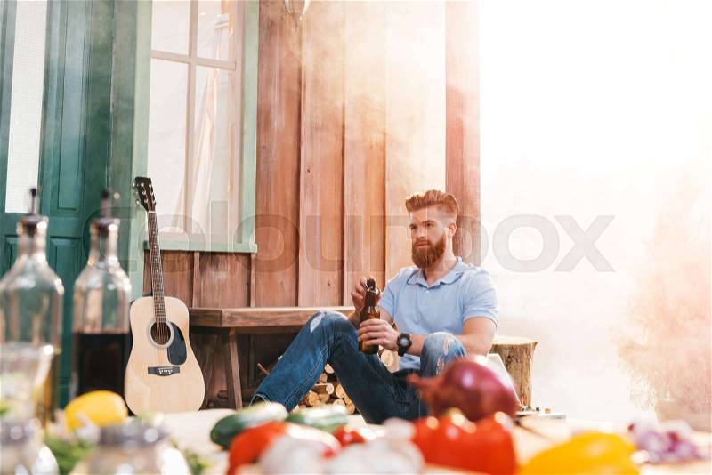 Handsome bearded young man sitting on porch and drinking beer , stock photo