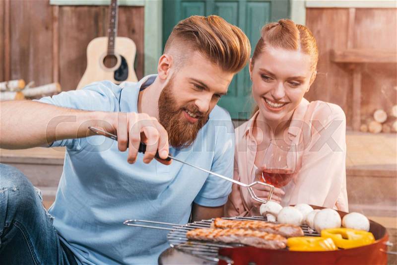 Man and woman roasting meat and vegetables on barbecue grill, woman holding wineglass, stock photo