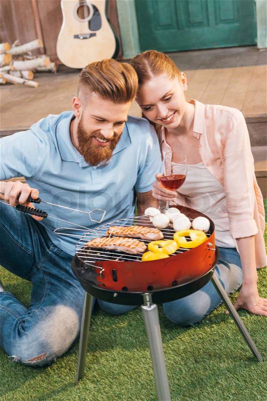 Man and woman roasting meat and vegetables on barbecue grill, woman holding wineglass, stock photo
