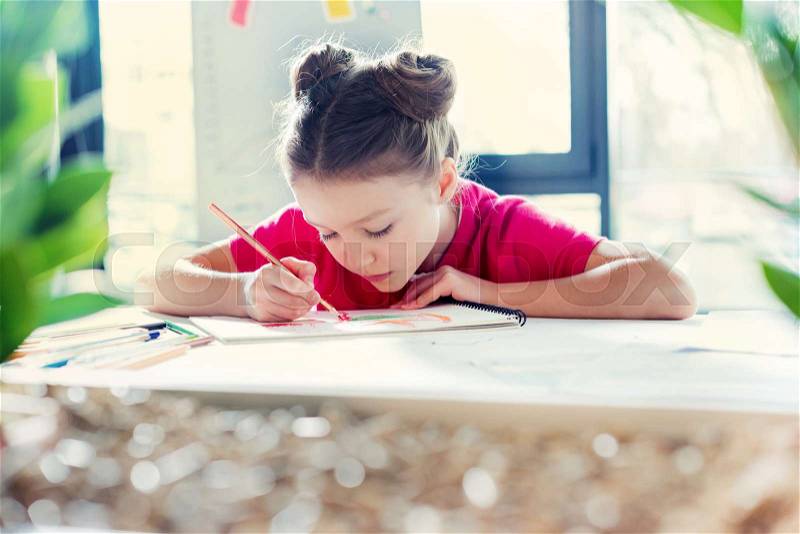 Concentrated little girl sitting at table and drawing with pencil, stock photo