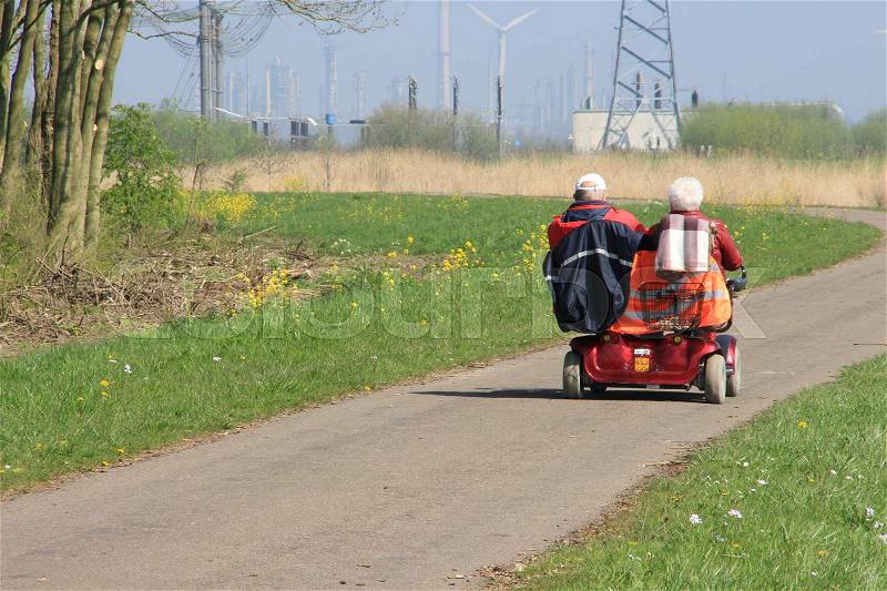 Retired couple, man and wife in a mobility scooter for two persons are travelling through the country side on a sunny day in beautiful spring and in the distance the emerging industry, stock photo