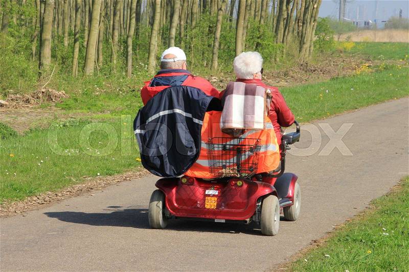 Retired couple, man and wife in a mobility scooter for two persons are travelling through the country side on a sunny day in beautiful spring and in the distance the emerging industry, stock photo