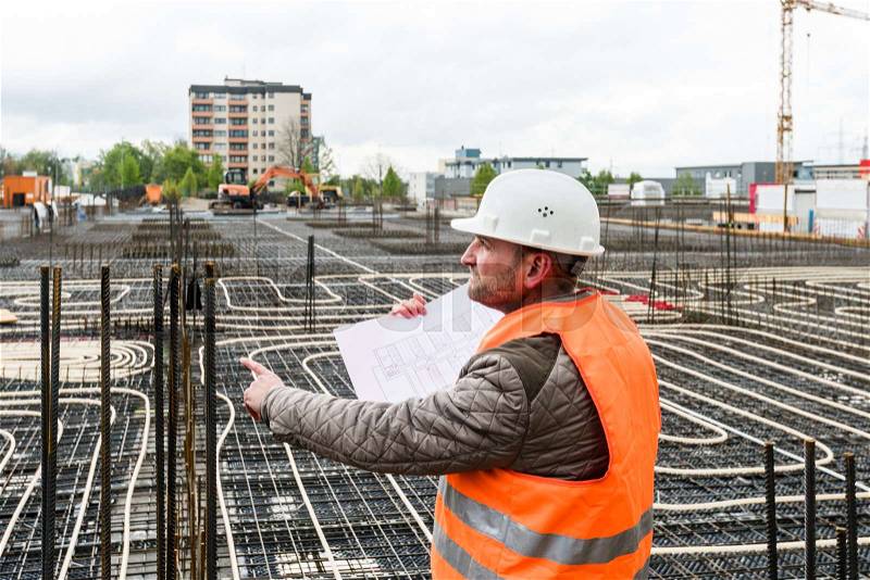 Construction supervisor oversees the construction works pointing with a plan in his hand, wearing a safety helmet and vest, stock photo