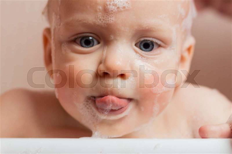 Close-up portrait of baby boy in soap foam in a bathtub. Big-eyed infant kid looking at camera and licking lips, stock photo