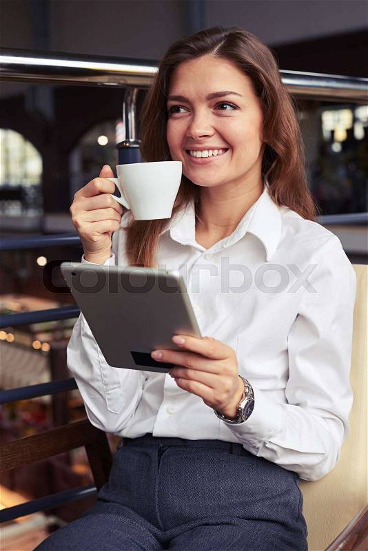 Vertical of gorgeous smiling business woman enjoying tasty cup of coffee and reading latest news on tablet during break with copy space, stock photo