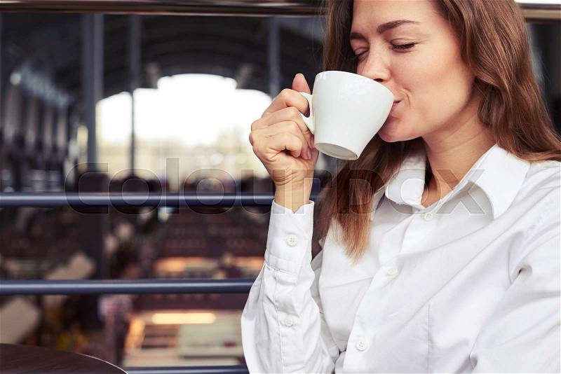 Close-up of very nice woman in white shirt tasting cup of espresso in café, stock photo