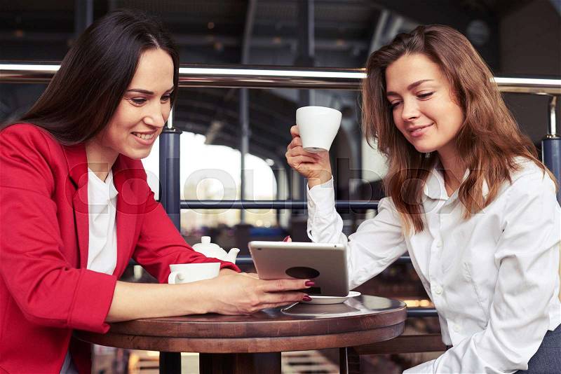 Close-up of two attractive charming colleagues chatting and photos on tablet during lunch with cup of coffee, stock photo