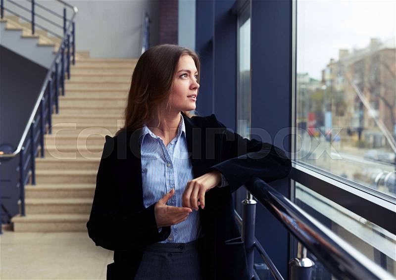 Close-up of charmed woman in black coat looking through the window. Office worker wearing formal clothes indoors , stock photo