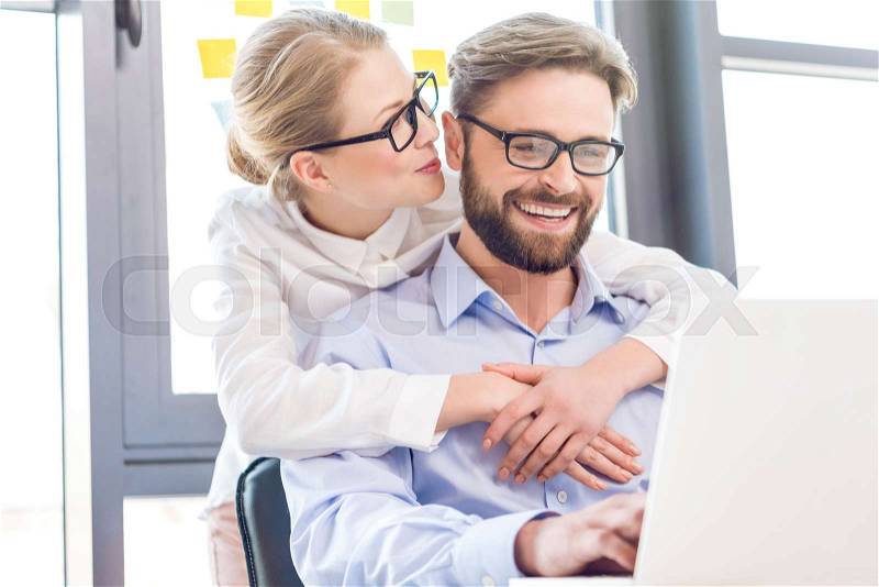 Businesswoman and businessman with eyeglasses hugging and working with laptop in office, stock photo