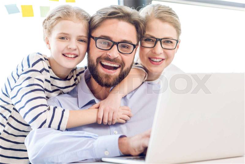 Happy father, mother and daughter hugging and using laptop in office, stock photo