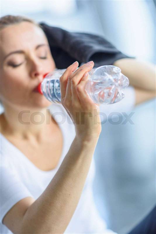 Tired sportswoman drinking water from sport bottle in sports center, stock photo