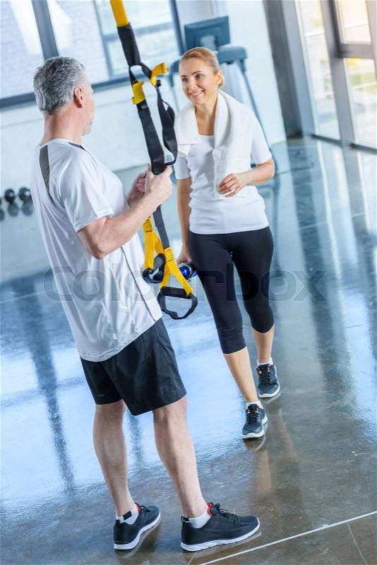 Sportswoman holding sport bottle and senior sportsman with resistance band talking in sports center, stock photo