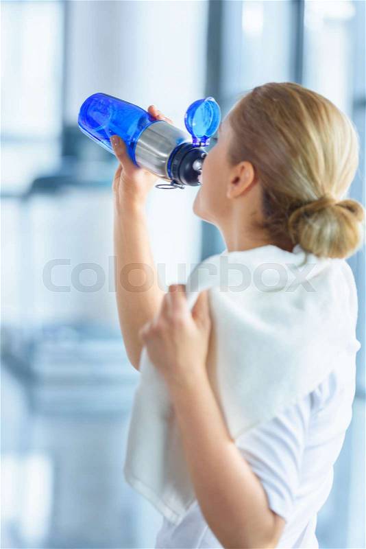 Sportswoman with towel drinking water from sport bottle in sports center, stock photo