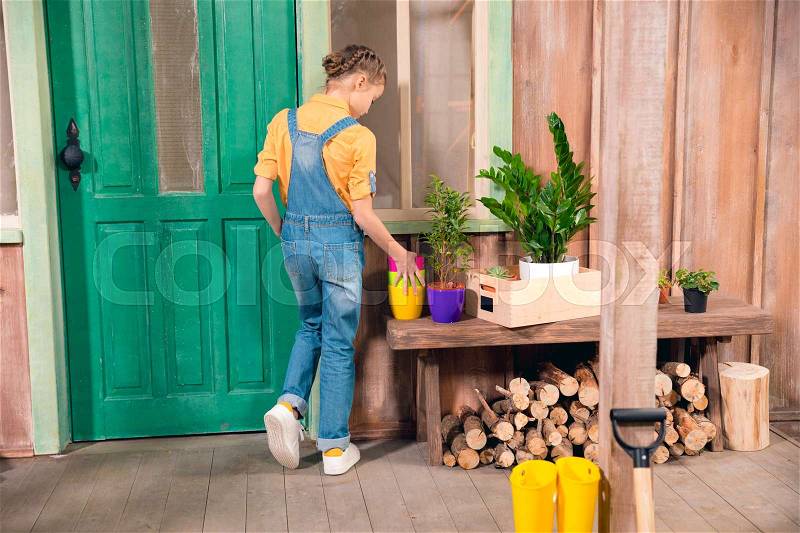 Back view of little girl in denim overalls standing on porch with flower pots, stock photo