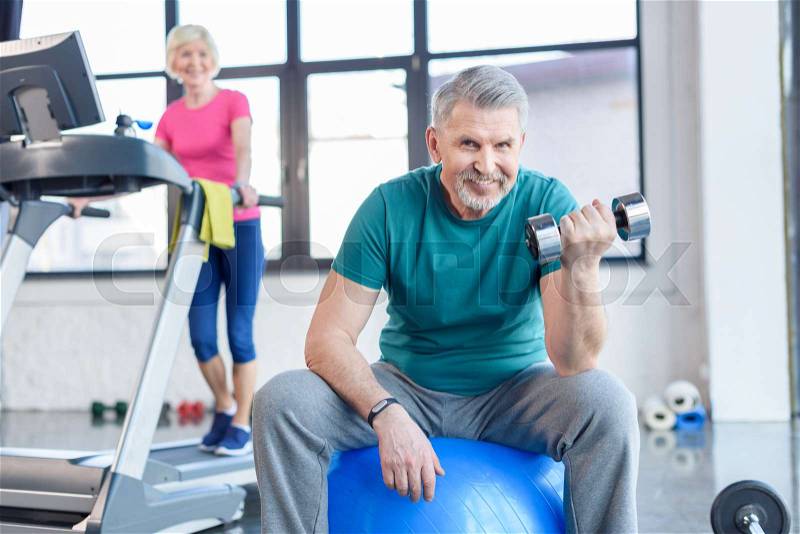 Senior sportsman sitting on fitness ball and training with dumbbell, sportswoman on treadmill behind. senior fitness class concept, stock photo