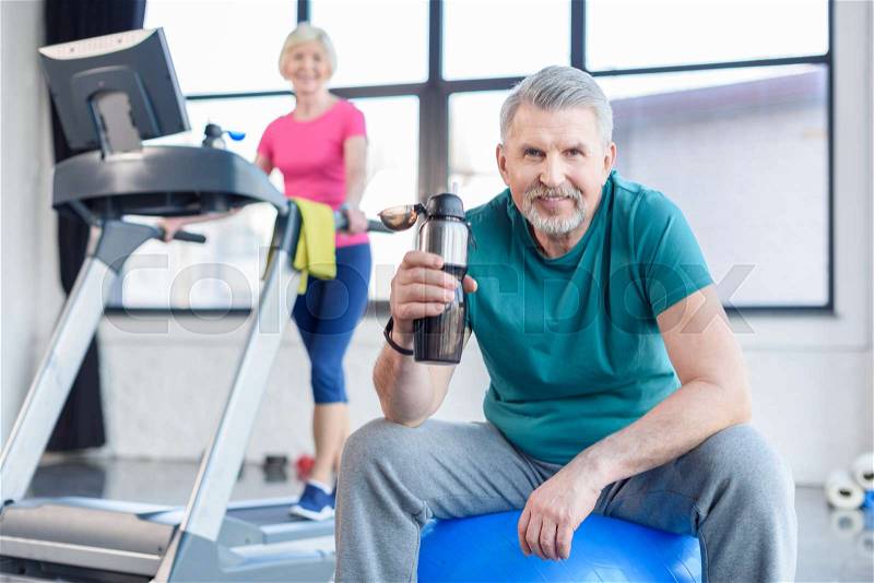 Senior sportsman sitting on fitness ball with sport bottle, sportswoman on treadmill behind. senior fitness class concept , stock photo