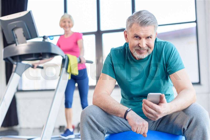 Senior sportsman sitting on fitness ball and using smartphone, sportswoman on treadmill behind. senior fitness class concept, stock photo