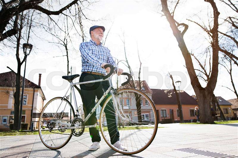 Handsome senior man in blue checked shirt with bicycle in town, looking up. Sunny spring day, stock photo