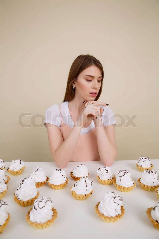 Vertical image of sensual woman sitting by the table with many cakes while looking away over cream background, stock photo
