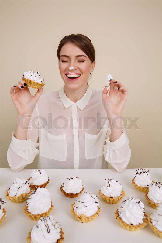 Cheerful funny young woman eating cakes and laughing over white background, stock photo