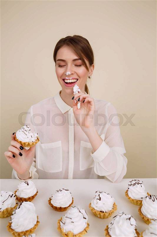 Happy playful young woman eating cupcakes at the table and having fun over white background, stock photo