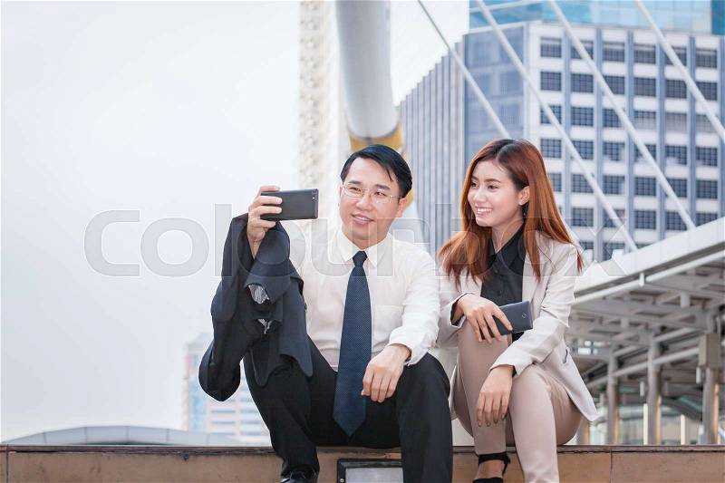 Businessman sitting and using phone to take photo as selfie with colleague together and modern office building background, stock photo